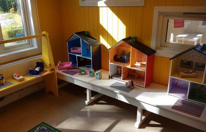 Berg barnehage inneområde med tre dukkehus, benk og bord og solen som skinner inn gjennom vinduet
