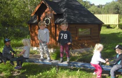 Bjerkaker barnehage uteområde med lekehus og seks barn på en huske en sommerdag med grønt gress
