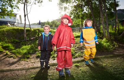 Tre barn med regntøy i Fjellvegen barnehage en sommerdag med grønt gress og skog
