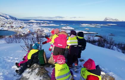Åtte barnehagebarn fra Sommarøy barnehage på tur på en liten fjelltopp en vinterdag med snø, blå himmel og med utsikt over fjell og hav