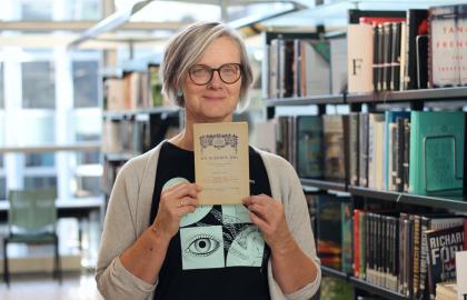 Kvinne med briller og grå cardigan holder opp en gammel bok og smiler mot kamera. Foto