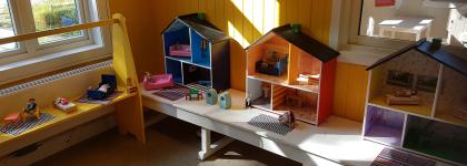 Berg barnehage inneområde med tre dukkehus, benk og bord og solen som skinner inn gjennom vinduet